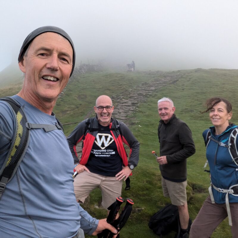 A photograph of four hikers on a hillside in the mist. They are smiling at the camera.