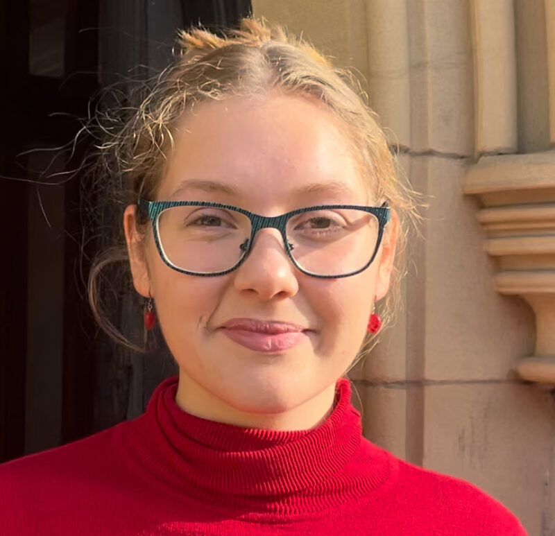 An image of a young woman, wearing a red top, dark rimmed glasses, with a pale stone column in the background