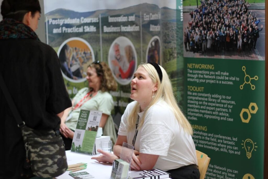 Photograph of Izabella at an event, she is sat at a table talking to someone. She is positioned at an angle to the camera so you cna see her side profile. She wears a white top and a black hairband is in her long bleached hair.