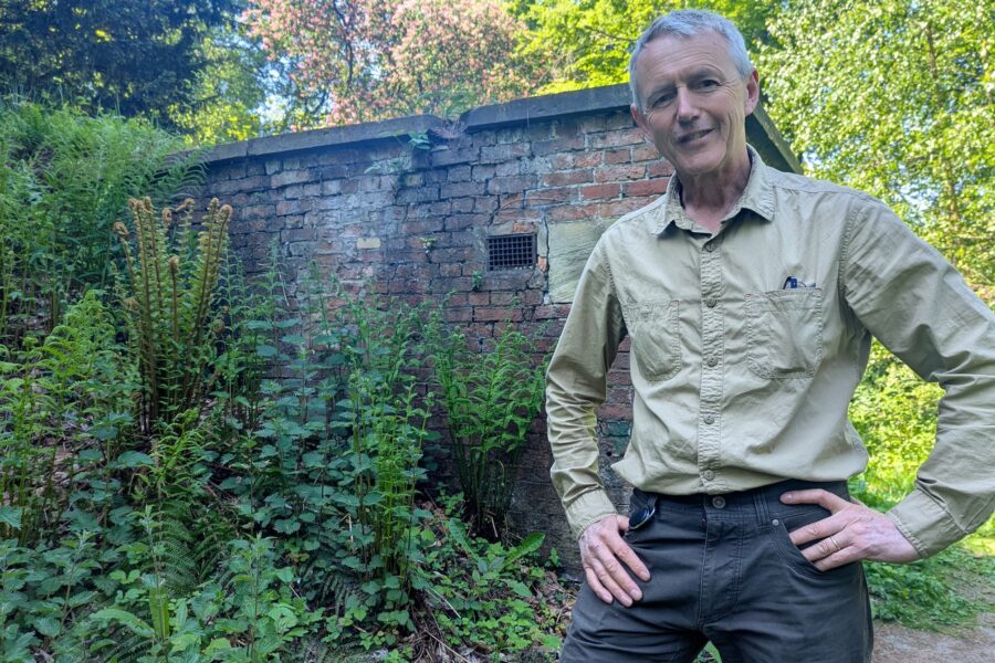 Simon Gill, Head of Heritage (Buildings and Landscapes) stands outside Wentworth Woodhouse's dilapidated Ice House. It is a small brick building surrounded by vegetation.