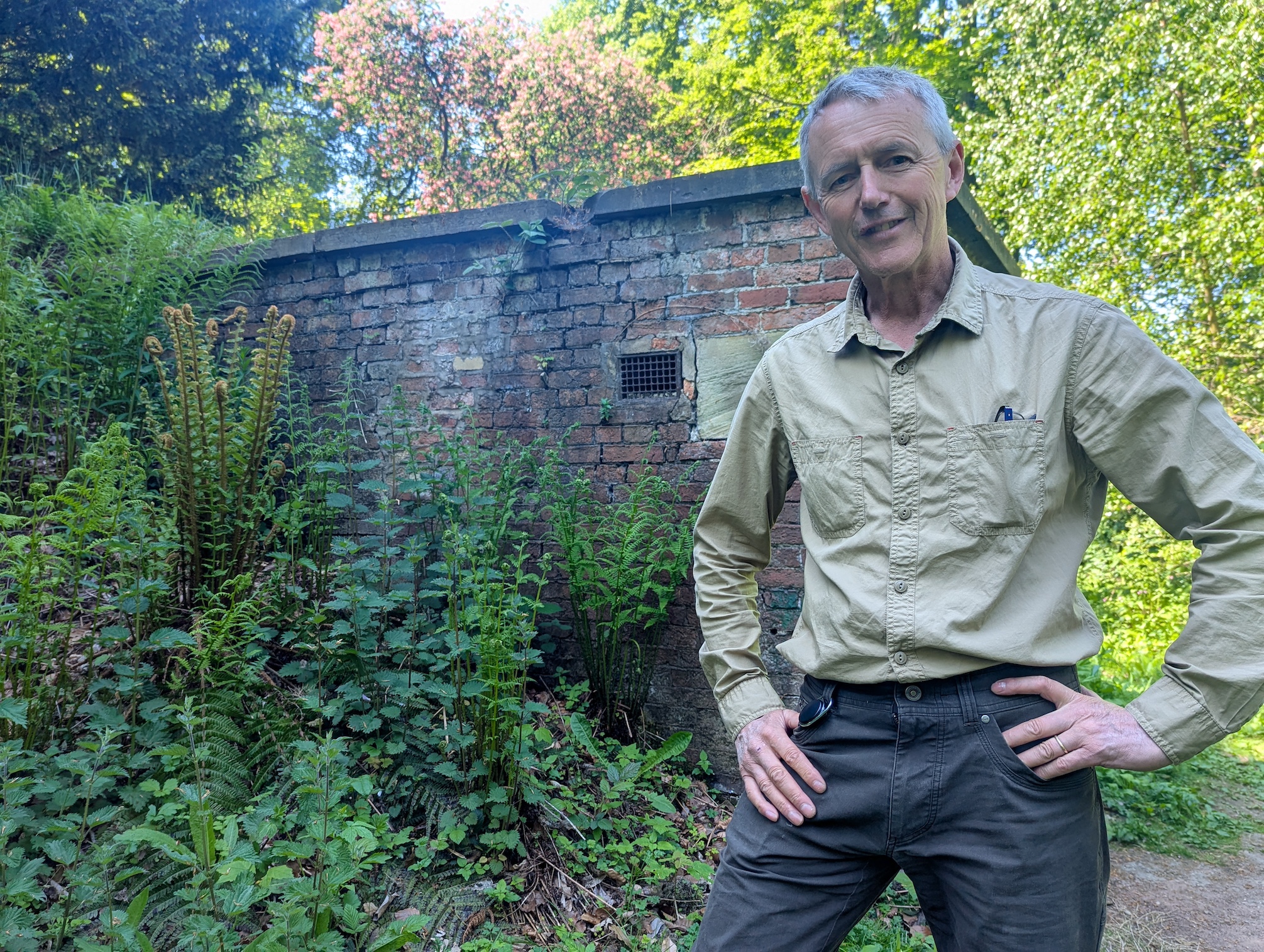 Simon Gill, Head of Heritage (Buildings and Landscapes) stands outside Wentworth Woodhouse's dilapidated Ice House. It is a small brick building surrounded by vegetation.