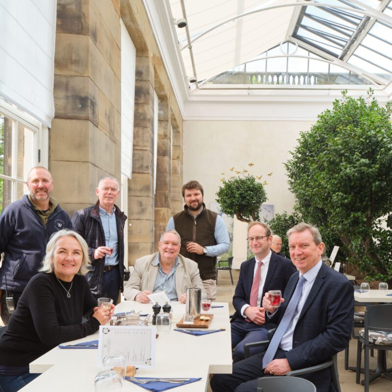 Seven people sitting around a table in a naturally brightly lit terrace. The terrace is part of the renovated Camellia House.