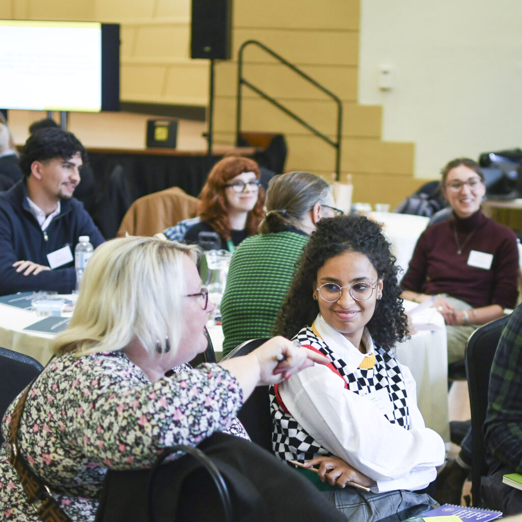 A group of people sit around tables and talk, everyone is smiling and looks relaxed and engaged.
