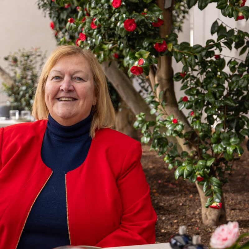 Dame Julie Kenny smiling while seated under a blooming rose bush.