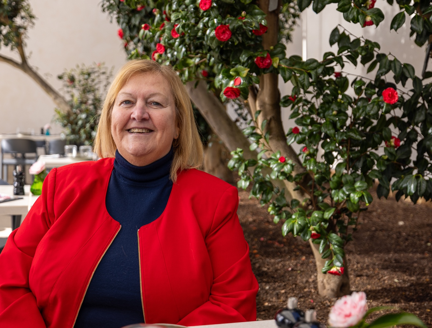 Dame Julie Kenny smiling while seated under a blooming rose bush.