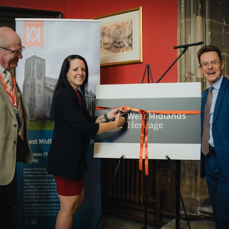 Three people stand around a sign that reads 'West Midlands Heritage Trust'. The sign has an orange ribbon tied around it, and one person is cutting the ribbon as part of an opening ceremony.