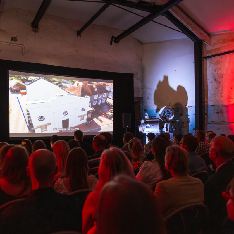 A photo of a full auditorium at the Selsey Pavillion Premiere, the crowd are sitting in dim, red lighting, watching a film on the big screen.