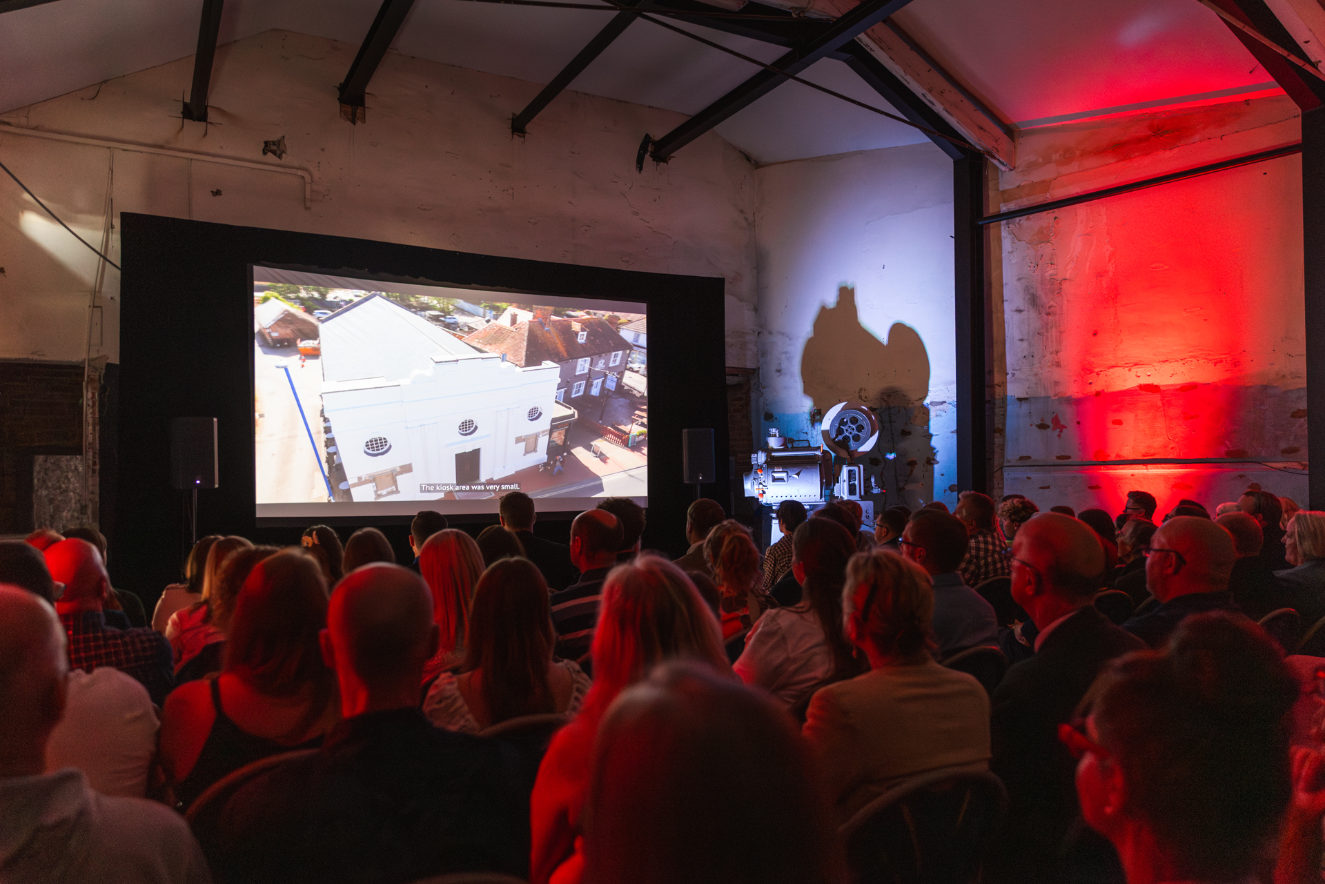 A photo of a full auditorium at the Selsey Pavillion Premiere, the crowd are sitting in dim, red lighting, watching a film on the big screen.