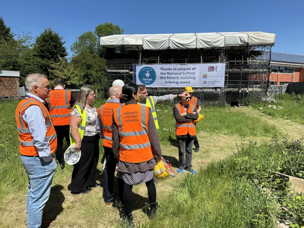 A group of people in hi-viz jackets stand on grass in front of a building covered in scaffolding.