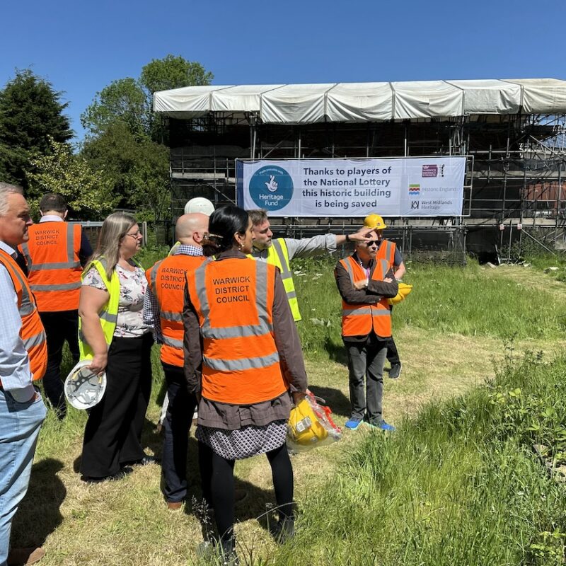 A group of people in hi-viz jackets stand on grass in front of a building covered in scaffolding.