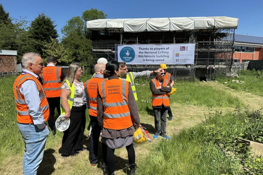 A group of people in hi-viz jackets stand on grass in front of a building covered in scaffolding.