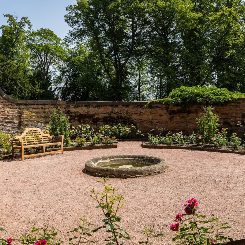 Photograph of the sensory rose garden. It's a circular space with flower beds filled with roses around a circular gravel area which has a stone circle at the centre.