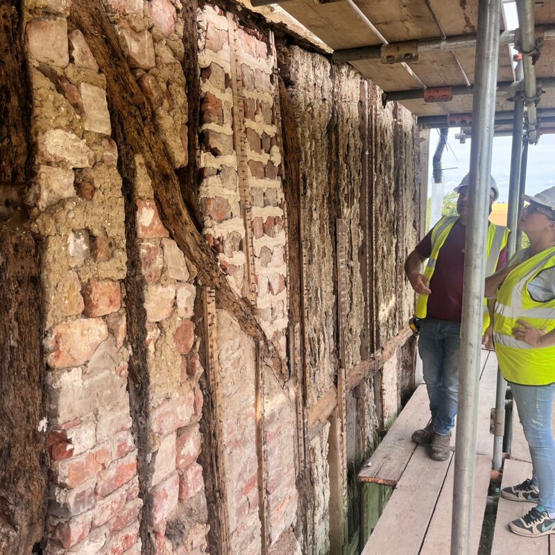 Two people in hi-viz jackets and hard hats stand on scaffolding inspecting the exposed bricks and timber of the chapel.