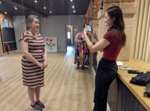 Eve, a young woman with long brown hair, is smiling and taking a video on her phone of another woman with grey hair and a brown and white striped dress.