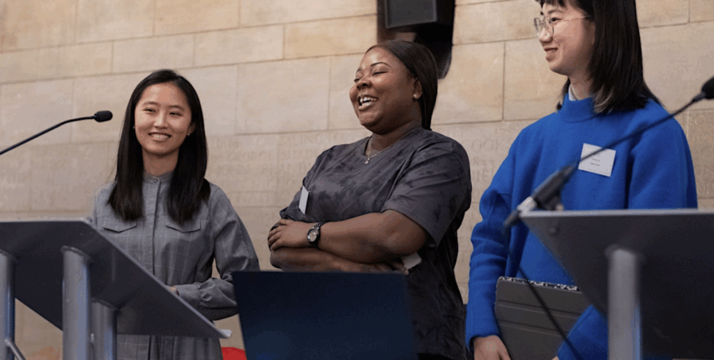 three young people stand on a stage behind a podium, they are smiling and giving a speech