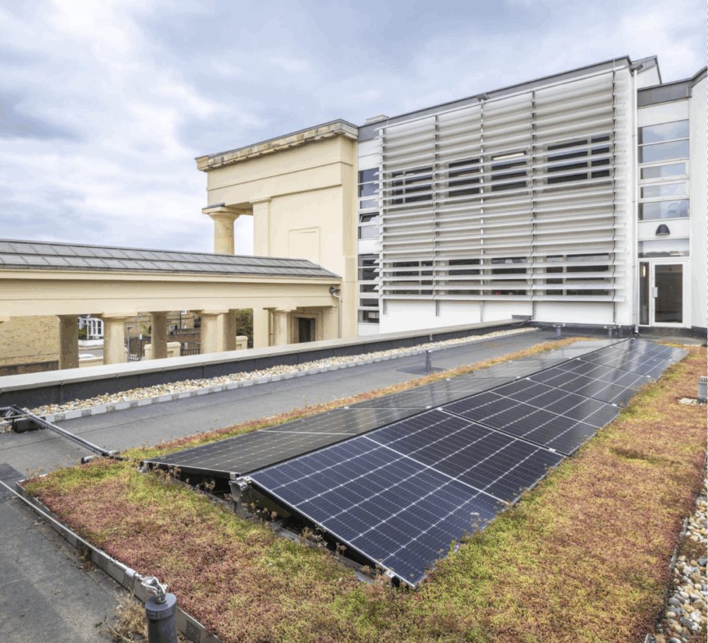 Solar panels on moss green roof of building, with taller section of building behind. In the background a building with cream columns joins the newer building.