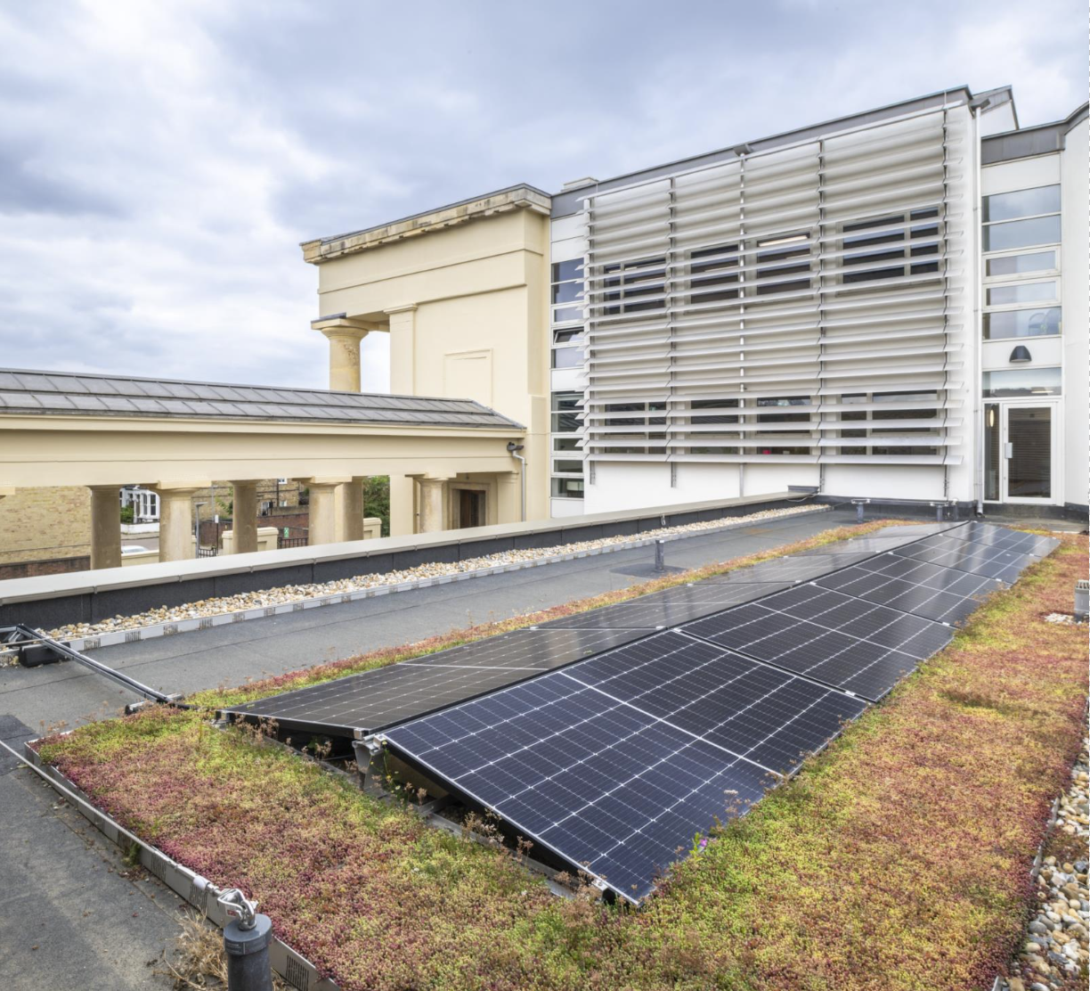 Solar panels on moss green roof of building, with taller section of building behind. In the background a building with cream columns joins the newer building.