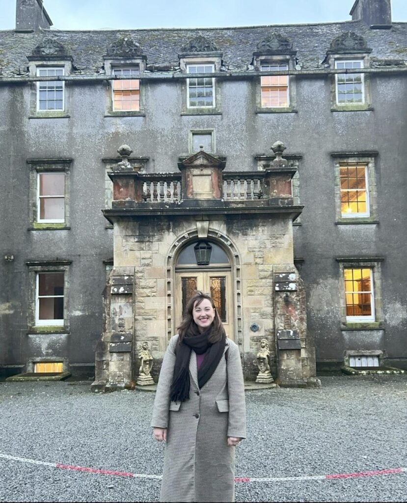 A dark haired lady in a full length coat stands in front of the house. There is a large entrance stone porch and the building is three storey with many windows.