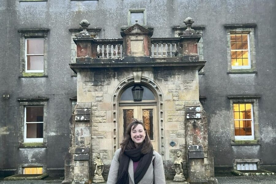 A dark haired lady in a full length coat stands in front of the house. There is a large entrance stone porch and the building is three storey with many windows.