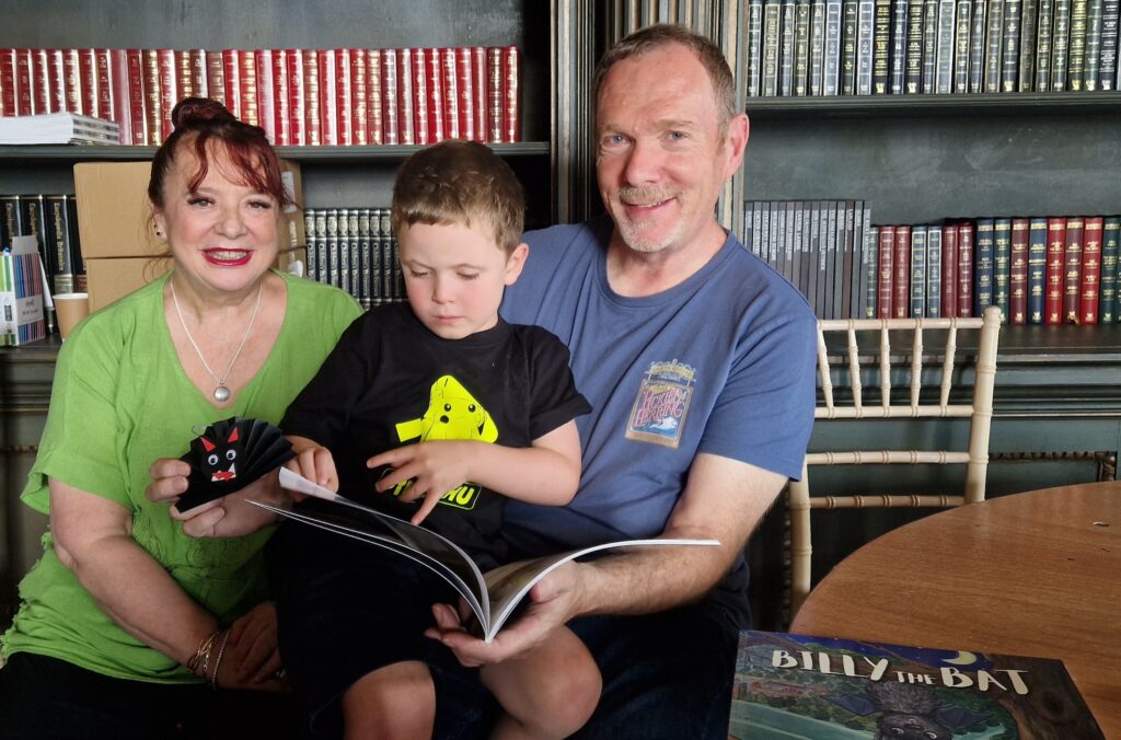 A man and woman sit in front of shelves of books. On their lap sits a young boy who is reading a large paperback book.