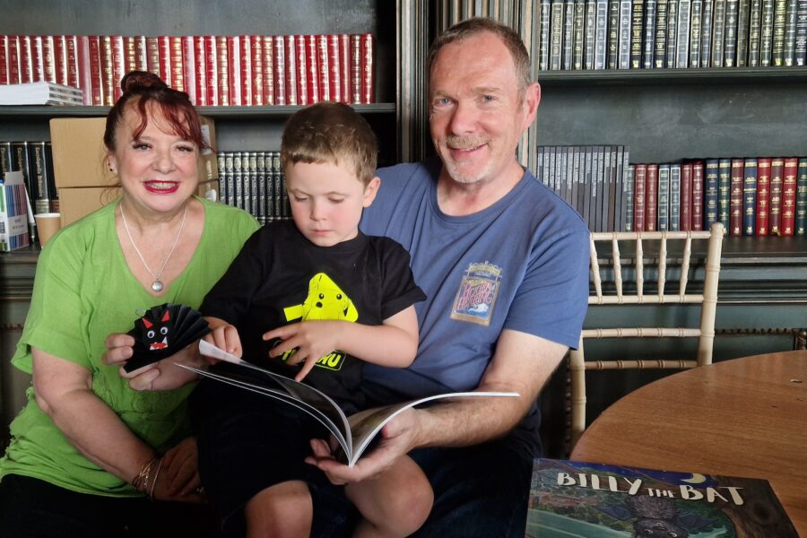 A man and woman sit in front of shelves of books. On their lap sits a young boy who is reading a large paperback book.