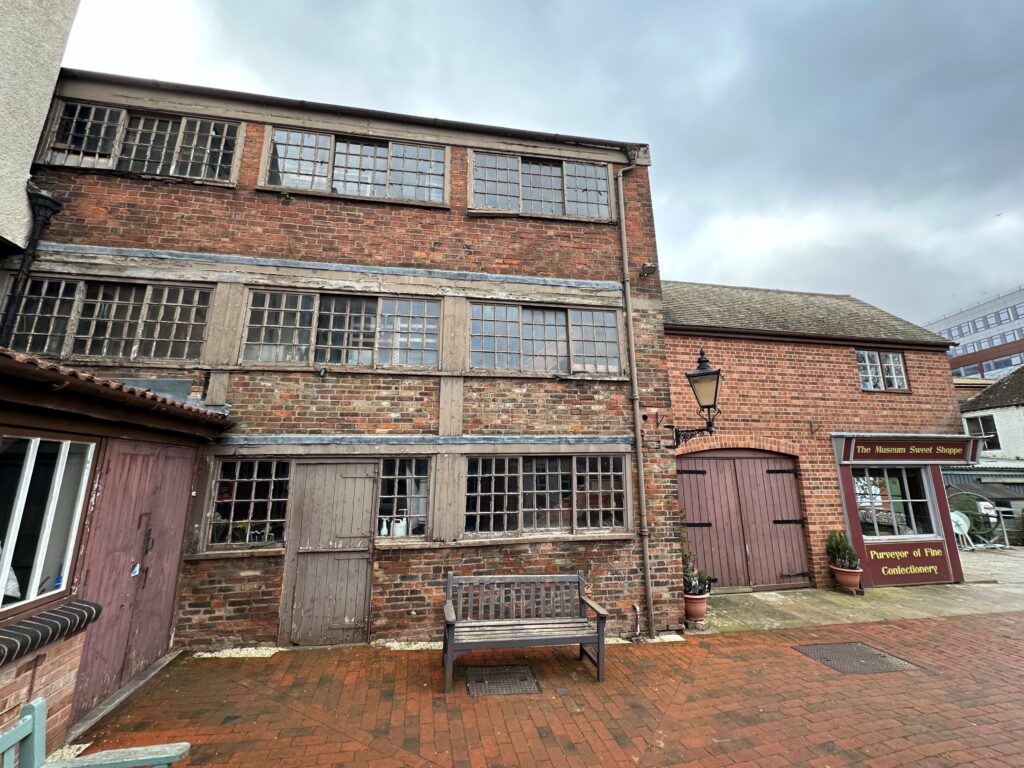 the pin factory, a red bricked three story building, it sits in a paved redbrick court yard and a bench site outside the building.