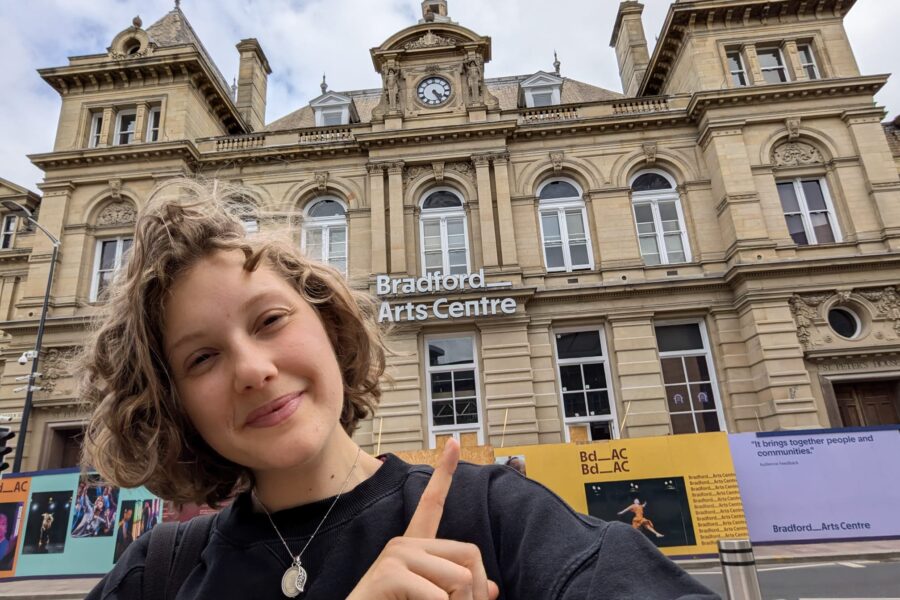 Sophie, a young white woman, is smiling and pointing towards the Bradford Art Centre building behind her.
