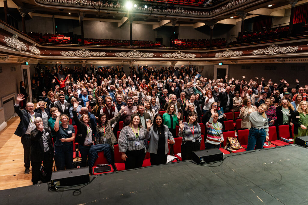 a large group of people in a theatre style setting waving to the camera.
