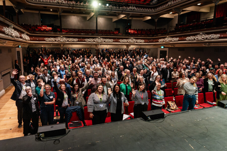 a large group of people in a theatre style setting waving to the camera.