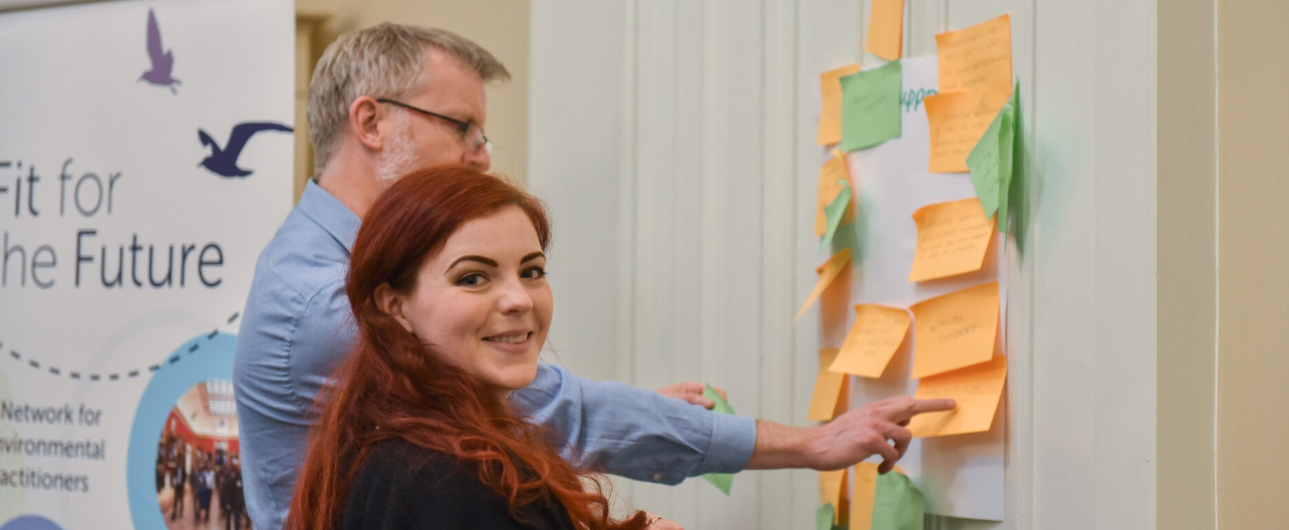 A young woman and a grey haired man are planning something using sticky notes on a wall. The young woman is looking towards the camera smiling.