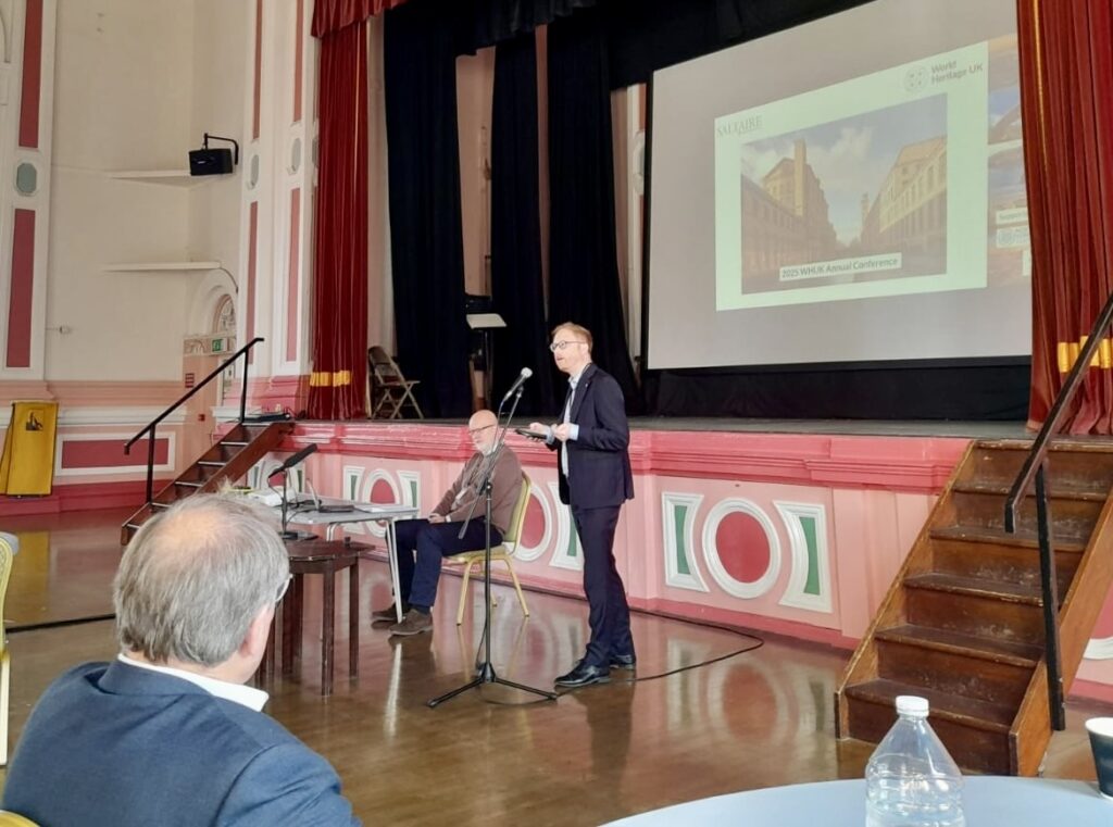 Jonathan Davies MP, a white man in a suit, presents at the front of the main ballroom of Victoria Hall in Saltaire. The room's walls have pink and green painted decoration, and there is a powerpoint slide behind Jonathan.