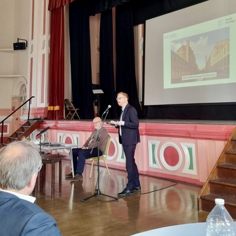 Jonathan Davies MP, a white man in a suit, presents at the front of the main ballroom of Victoria Hall in Saltaire. The room's walls have pink and green painted decoration, and there is a powerpoint slide behind Jonathan.