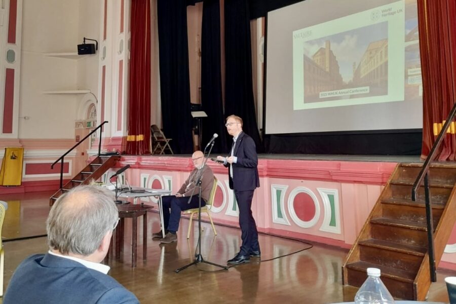 Jonathan Davies MP, a white man in a suit, presents at the front of the main ballroom of Victoria Hall in Saltaire. The room's walls have pink and green painted decoration, and there is a powerpoint slide behind Jonathan.