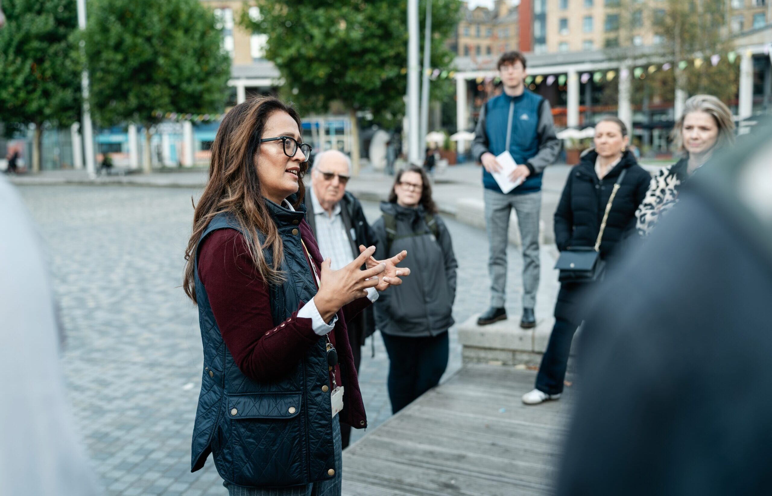 A group of people are looking at one person in the centre of the image who is leading a walking tour. The background is grey street pavement.