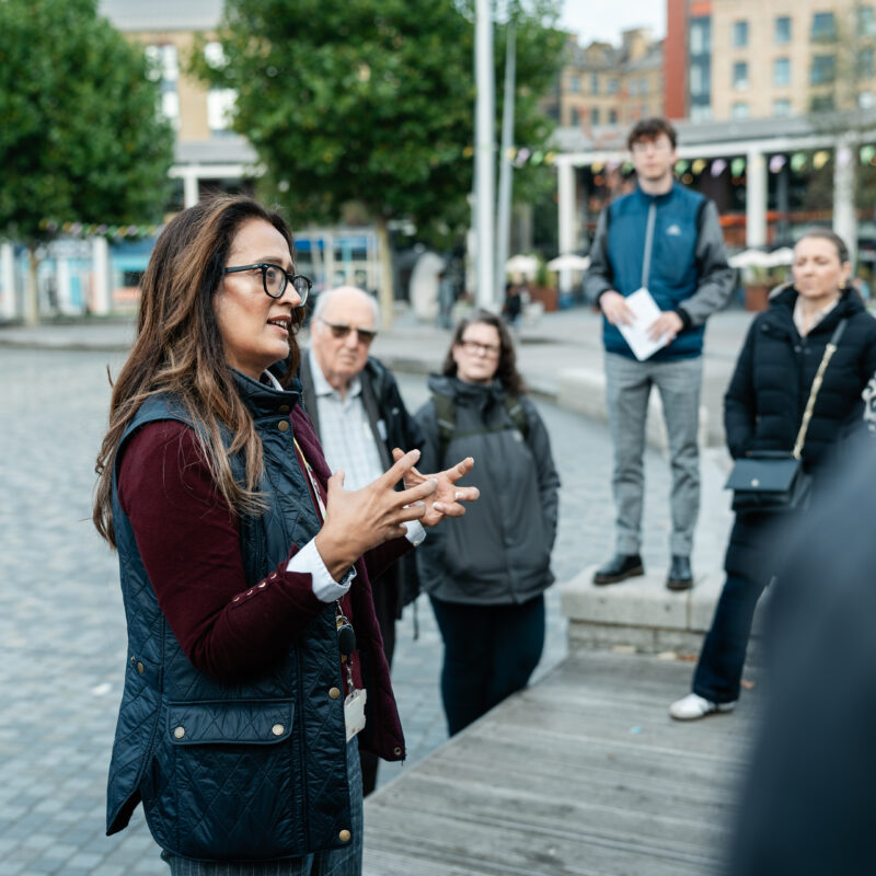 A group of people are standing outside, in a city centre. A tour guide is talking to the group.