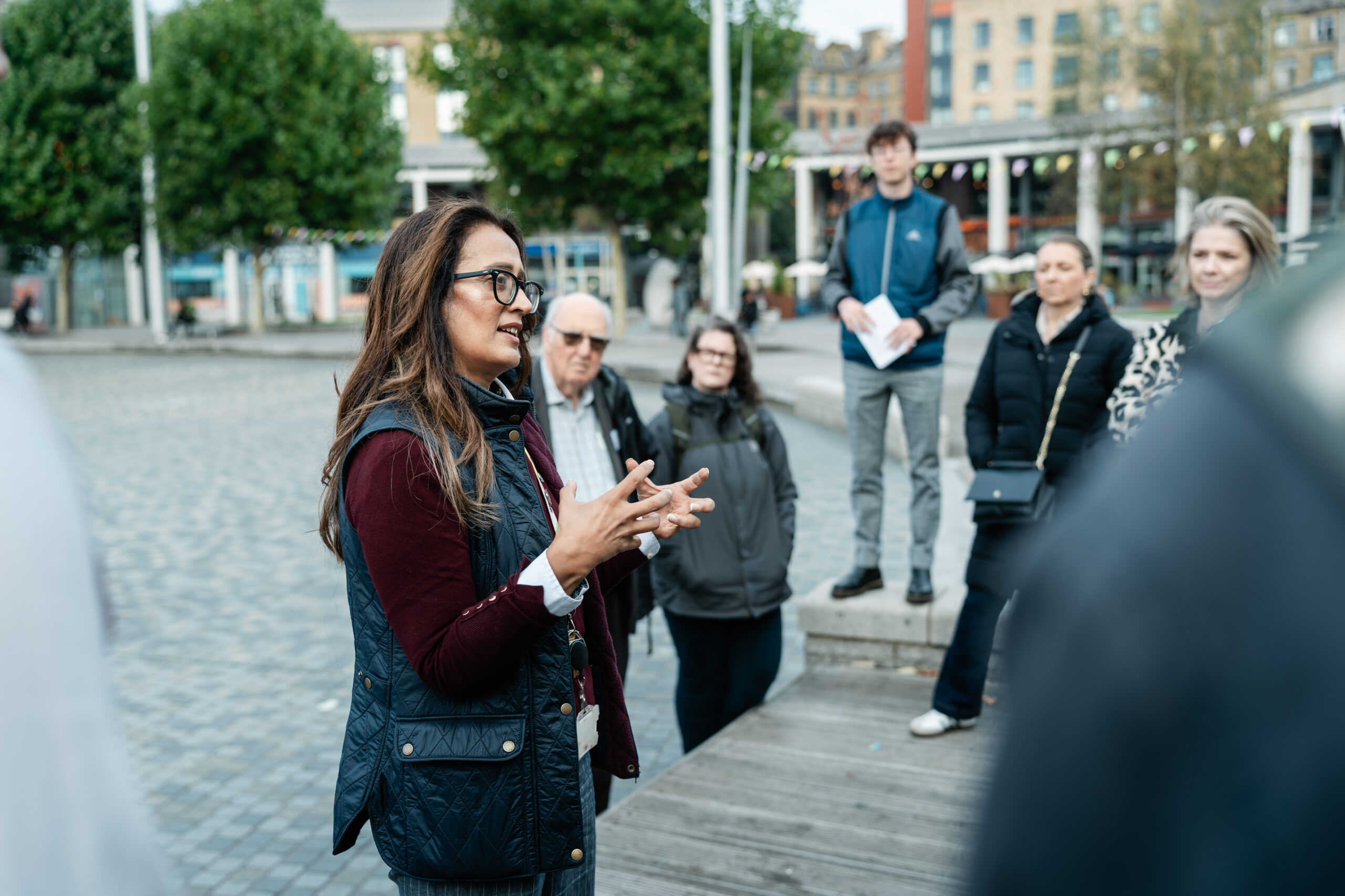 A group of people are standing outside, in a city centre. A tour guide is talking to the group.