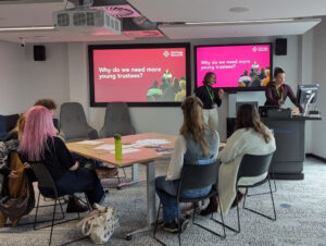 Two young women are leading a workshop. Other young people are sitting listening and looking at a screen that says 'why do we need more young trustees?'