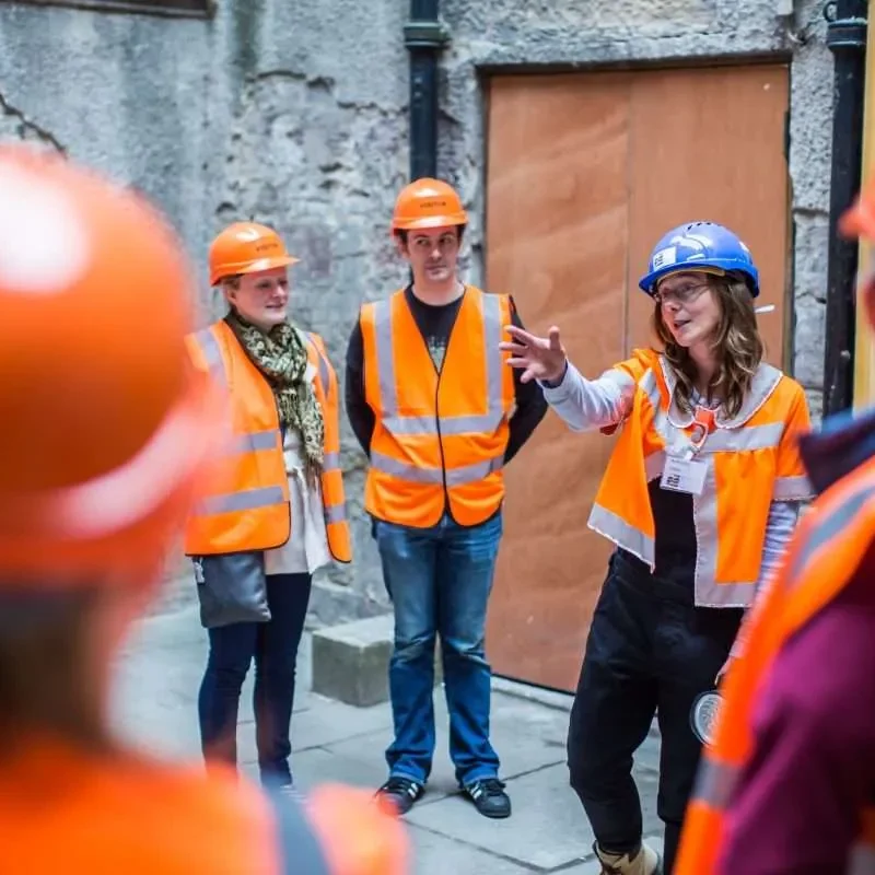 A group of people in orange high-vis vests and hard hats listen to a woman carrying a torch.