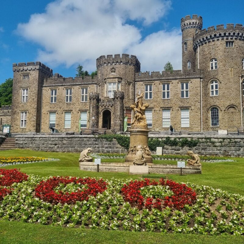 Cyfarthfa Castle with a blue sky. A flowerbed in the foreground with flowers in the shape of 200.