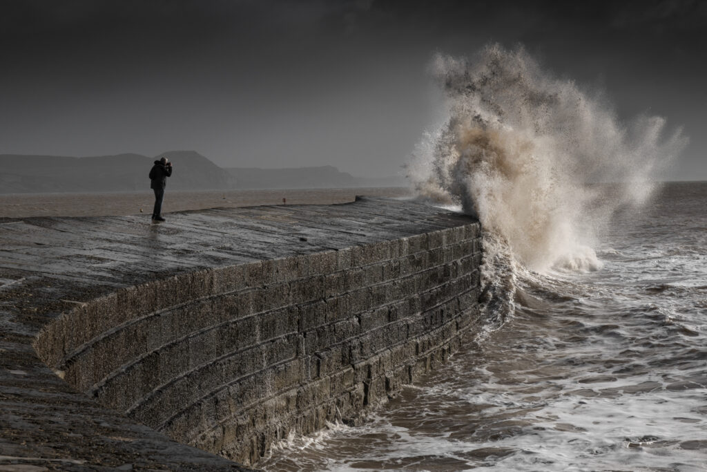 Large white wave crashing against a stone barrier on a headland, with a person in the mid ground taking a photograph of the wave