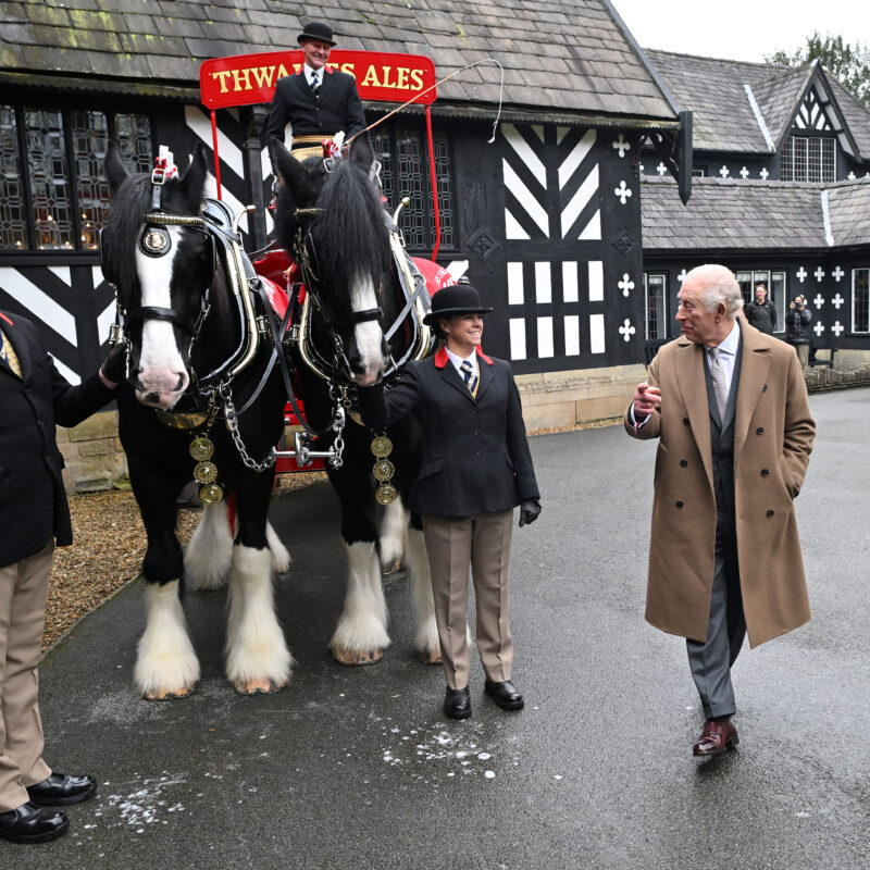 King Charles III stands outside Samlesbury Hall. Next to him is a cart and two shire horses and three people in black jackets and bowler hats supporting the horses.