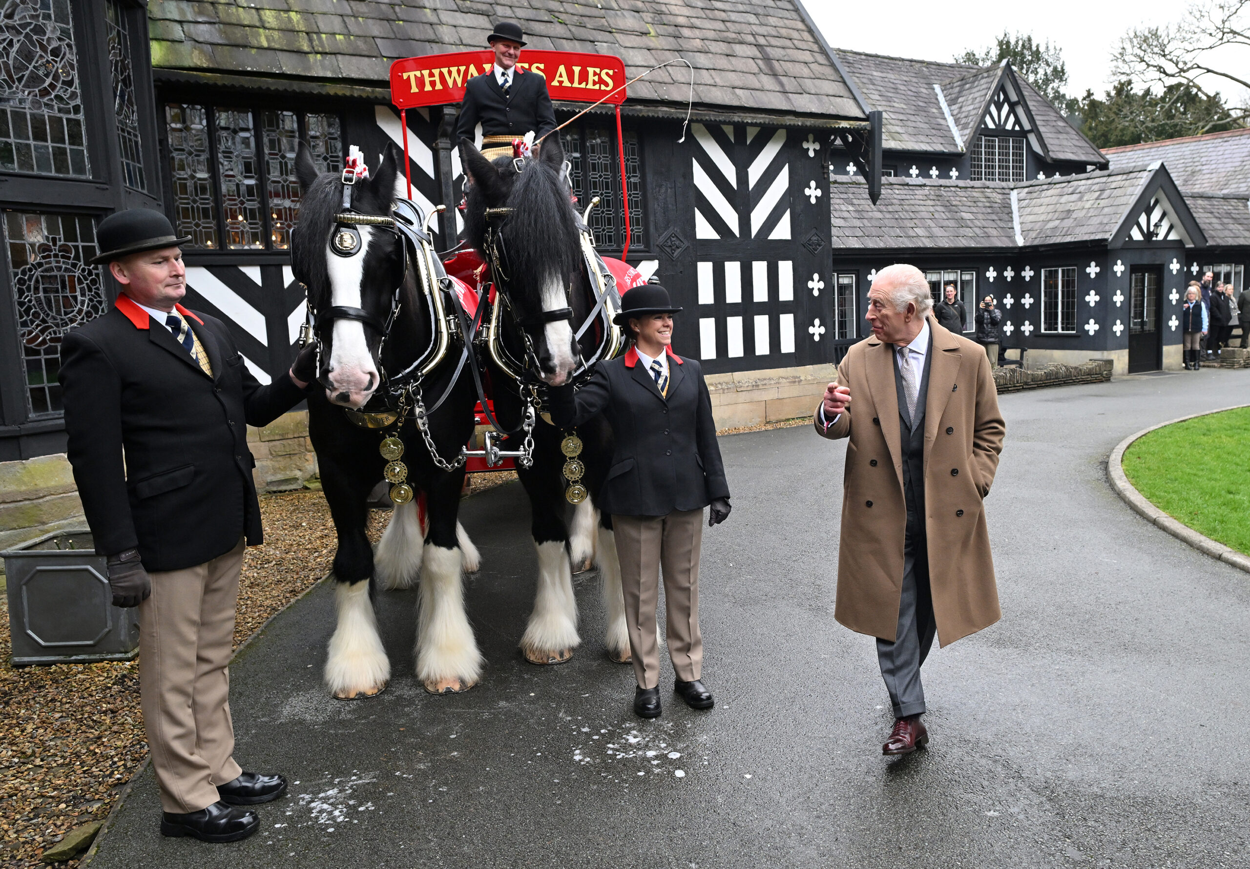 King Charles III stands outside Samlesbury Hall. Next to him is a cart and two shire horses and three people in black jackets and bowler hats supporting the horses.