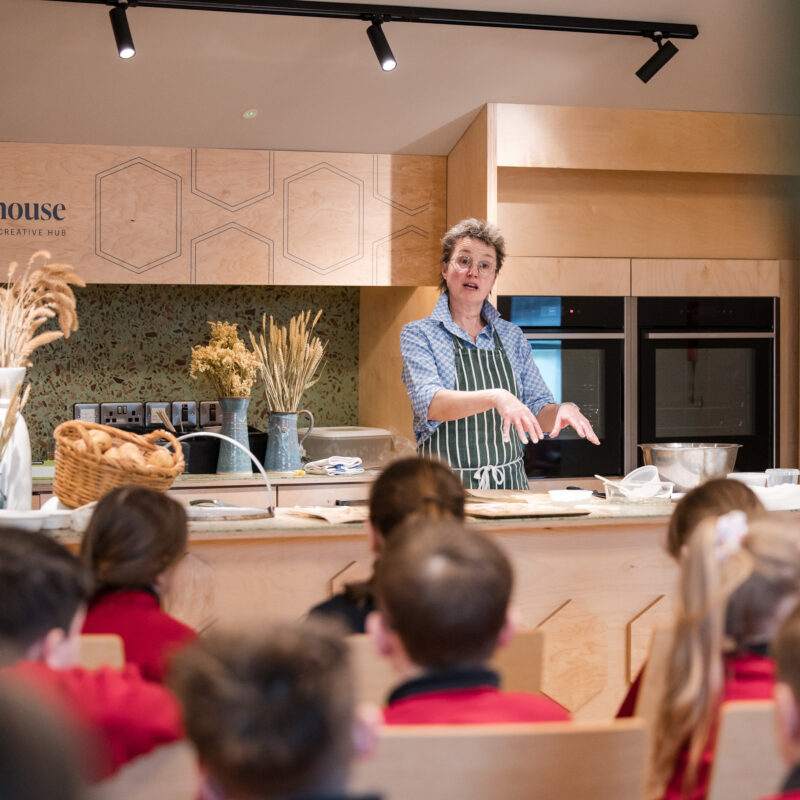 A white woman with short hair and glasses is facing a group of school children sitting on chairs. She is standing at a counter with cooking equipment, and sheaths of wheat in vases around her. The words 'The Courthouse shared space creative hub' are printed on the wall behind her.