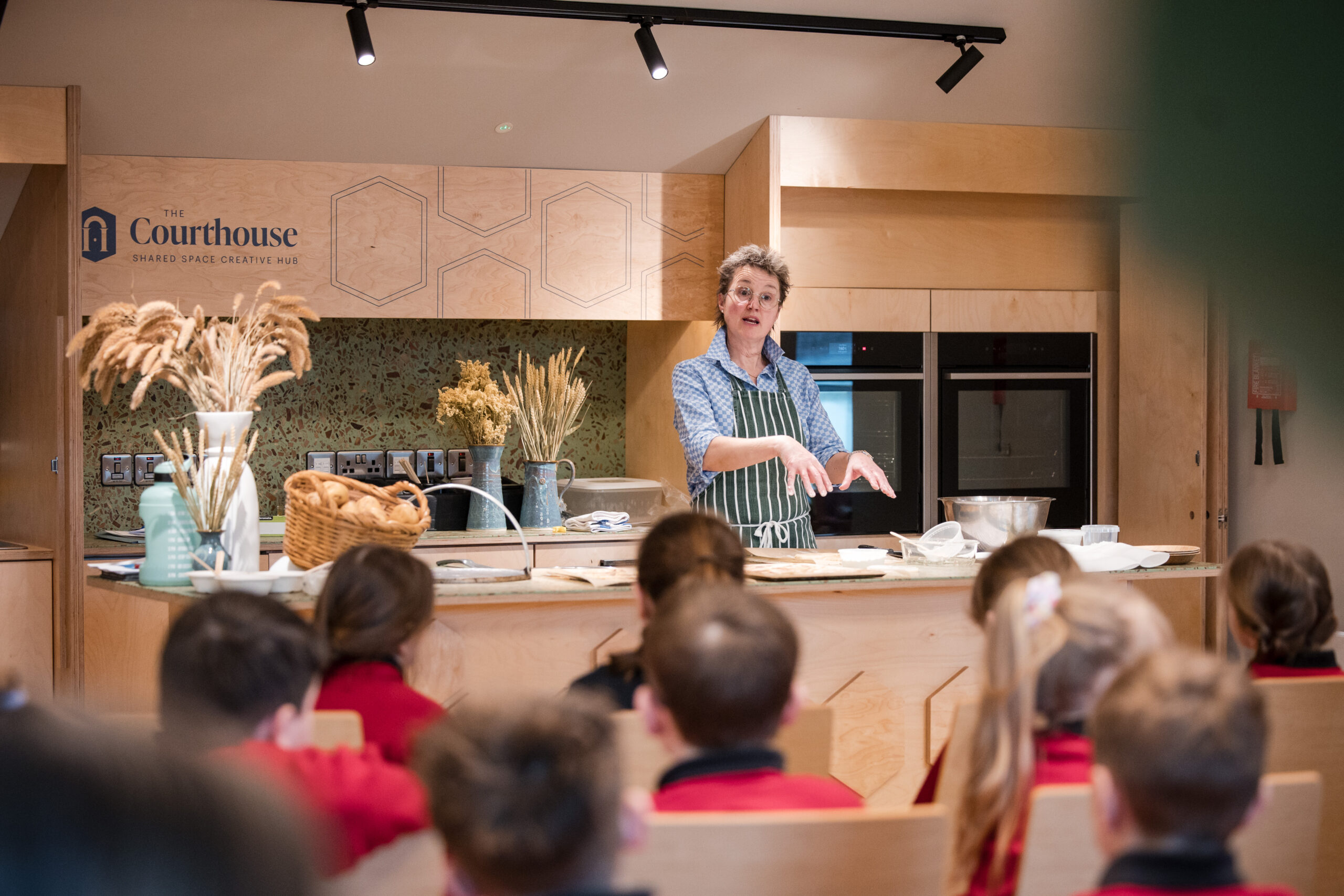 A white woman with short hair and glasses is facing a group of school children sitting on chairs. She is standing at a counter with cooking equipment, and sheaths of wheat in vases around her. The words 'The Courthouse shared space creative hub' are printed on the wall behind her.