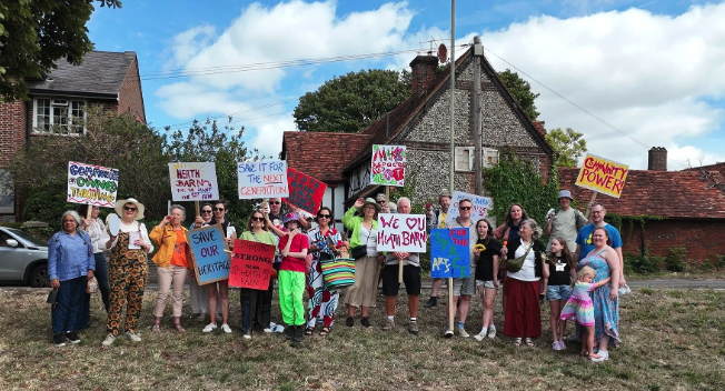 A group of people stand outside the barn. They hold placards saying things such as 'We love our barn' and 'Community power'.