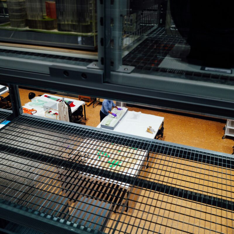 View through gap in museum storage shelves. Through the shelves, tables are visible. A member of staff wearing gloves is repacking an object into a storage box at one of the tables.