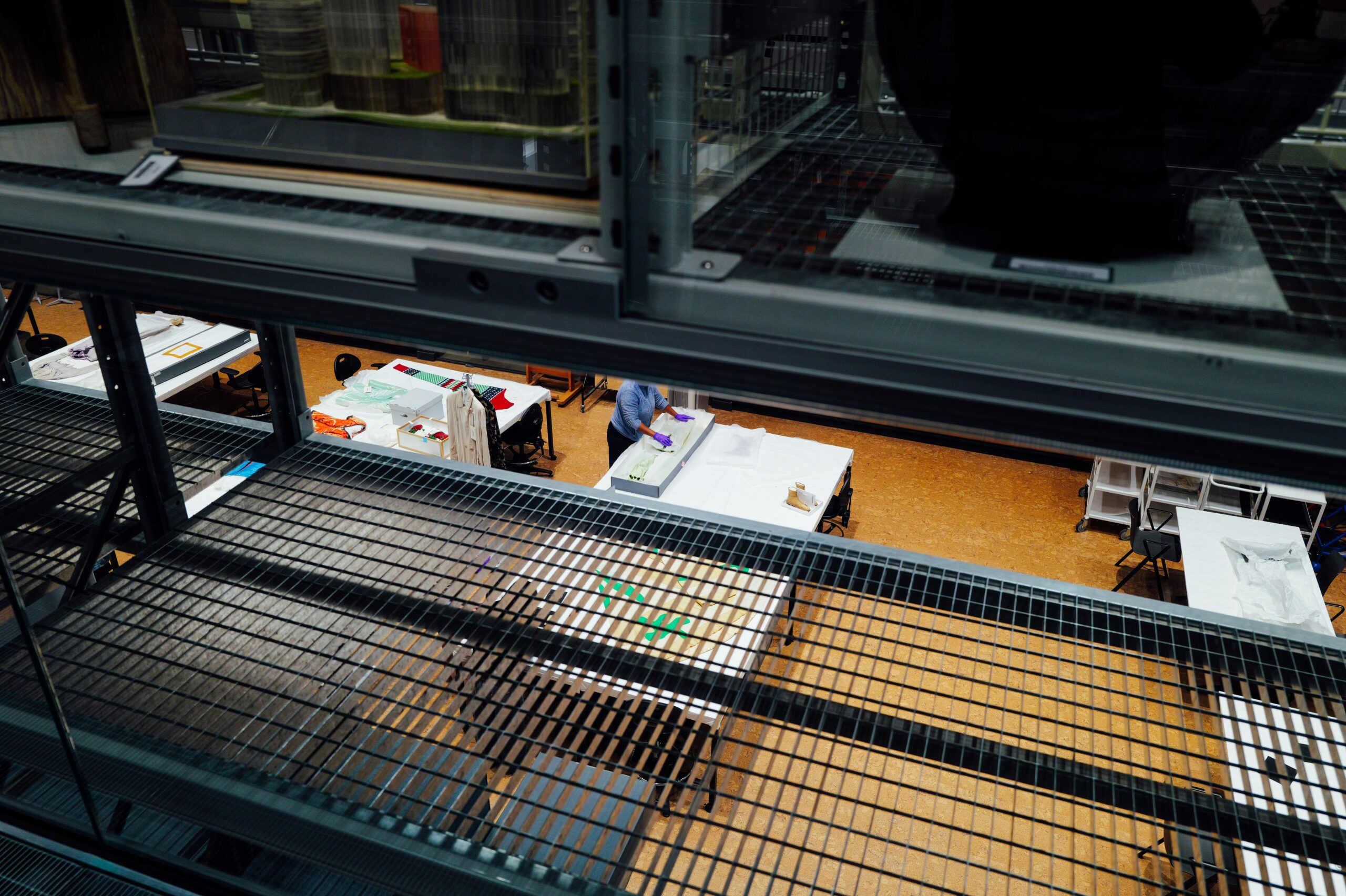View through gap in museum storage shelves. Through the shelves, tables are visible. A member of staff wearing gloves is repacking an object into a storage box at one of the tables.