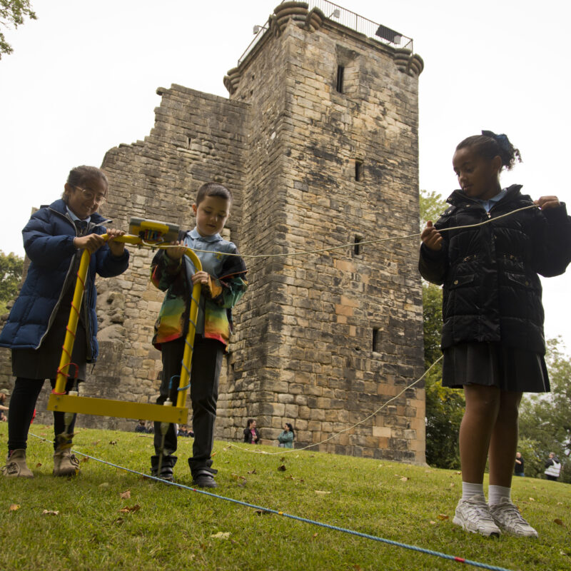 Three schoolchildren use specialist equipment to help survey the grounds around Crookston Castle, the ruins of which can be seen in the background.