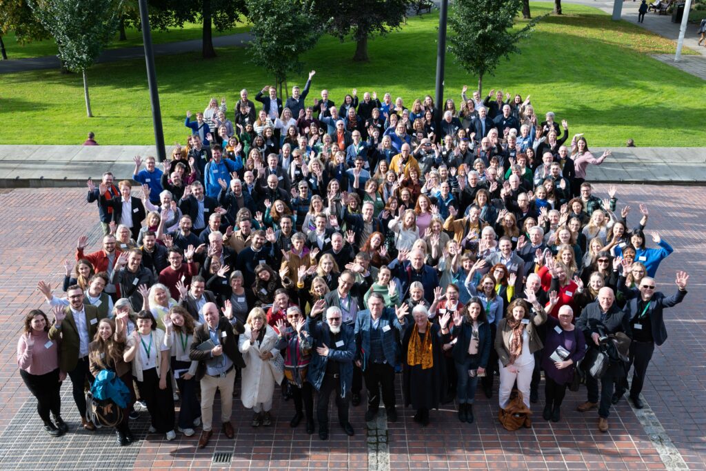 A photograph looking down on to a large group of people. They are all smiling, looking up at the camera and waving.