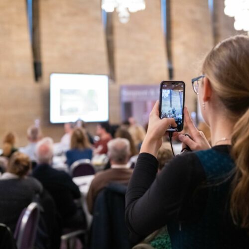shot from behind a young person who is filming the action on a stage at conference on her mobile phone